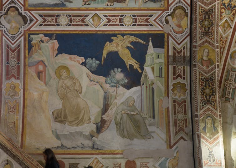 A fresco shows a haloed man kneeling in prayer as rays of light descend from a winged angel above, while a nun sits nearby reading from a book.