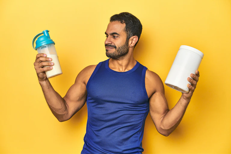 A fit man holds up a shake bottle with liquid in one hand and a white container in the other hand.