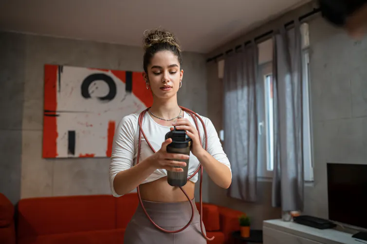 A young woman in gym clothes holds a supplement shaker bottle in her hands.