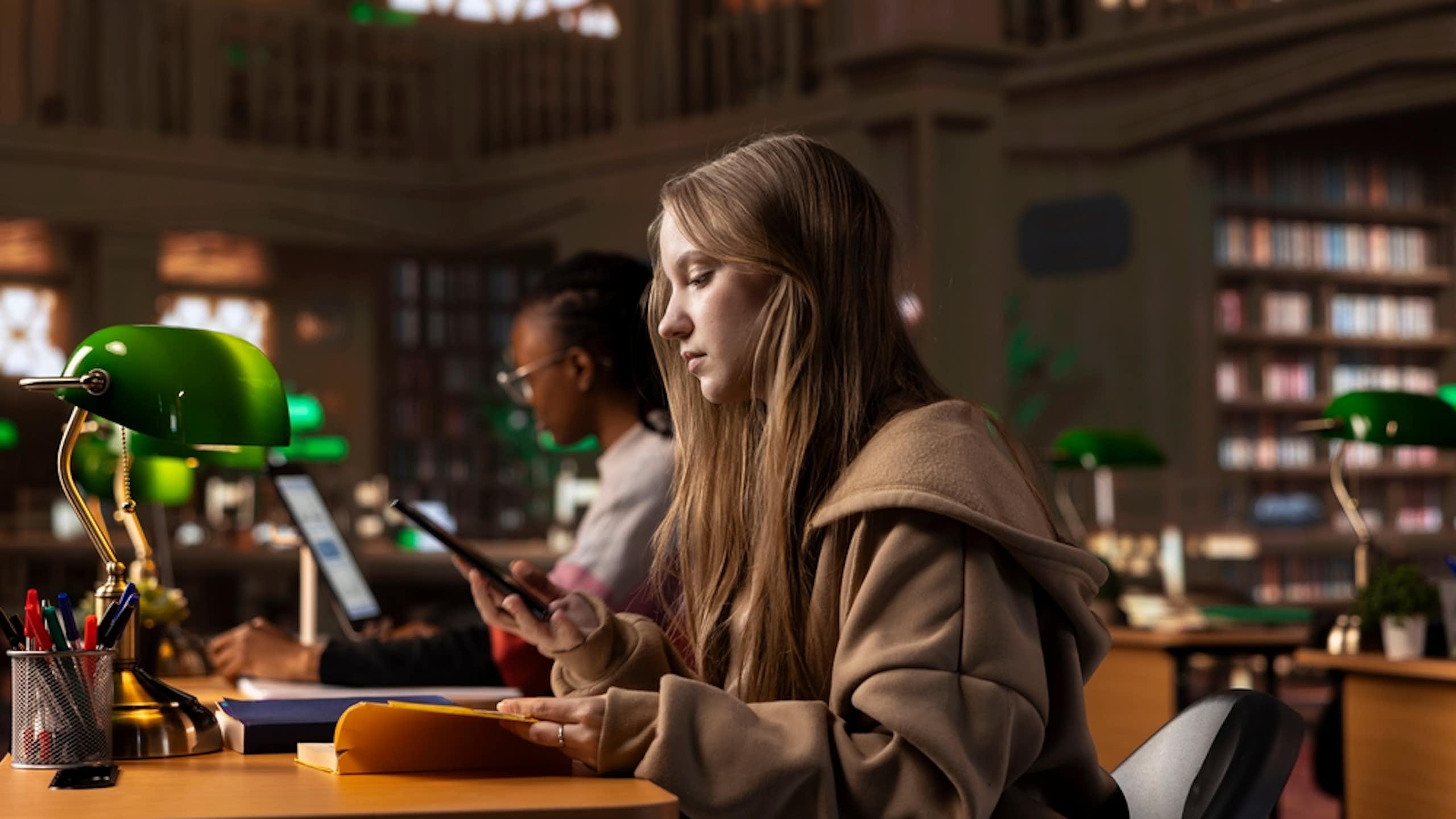 Une étudiante consultant un livre et prenant des notes sur son smartphone, en bibliothèque.