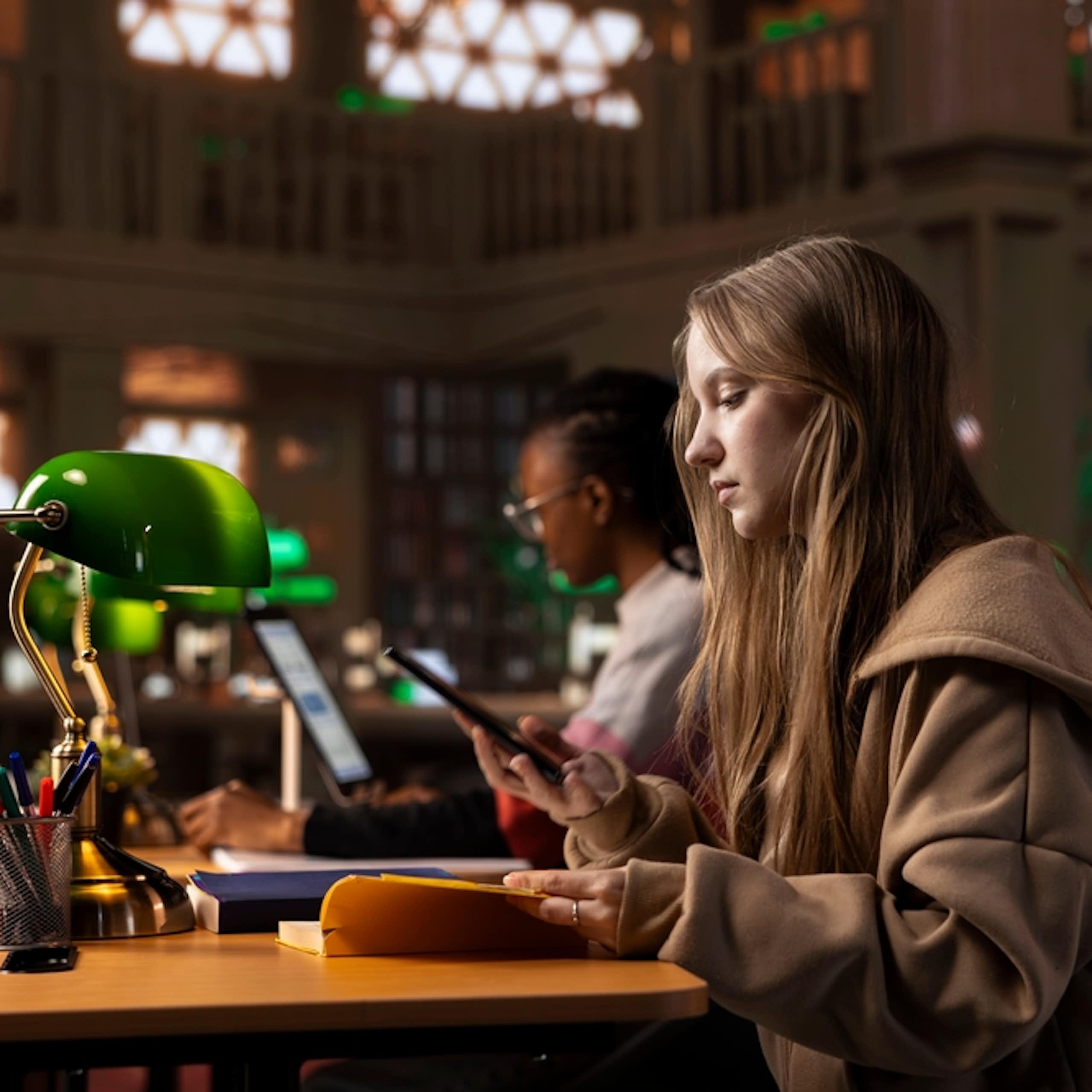 Une étudiante consultant un livre et prenant des notes sur son smartphone, en bibliothèque.