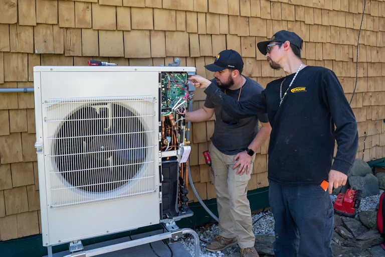 Two people stand next to a building and work on a square piece of equipment.