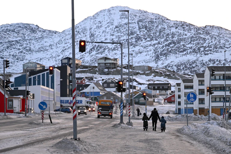 Three people walk through a town with a large snow-covered mountain in the background.