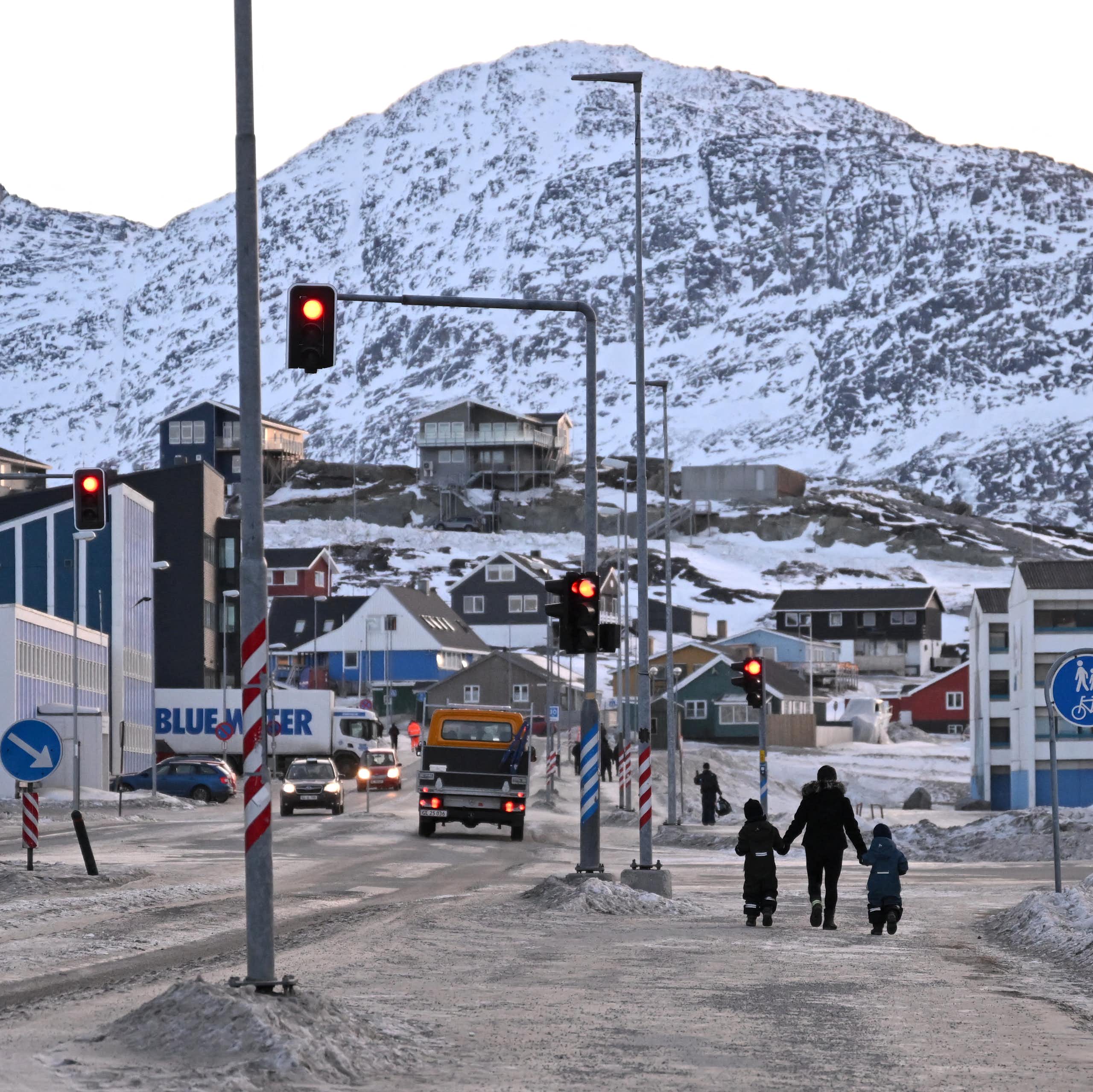 Three people walk through a town with a large snow-covered mountain in the background.