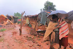 Two women stand outside in the rain with red-orange mud everywhere, and houses washed away