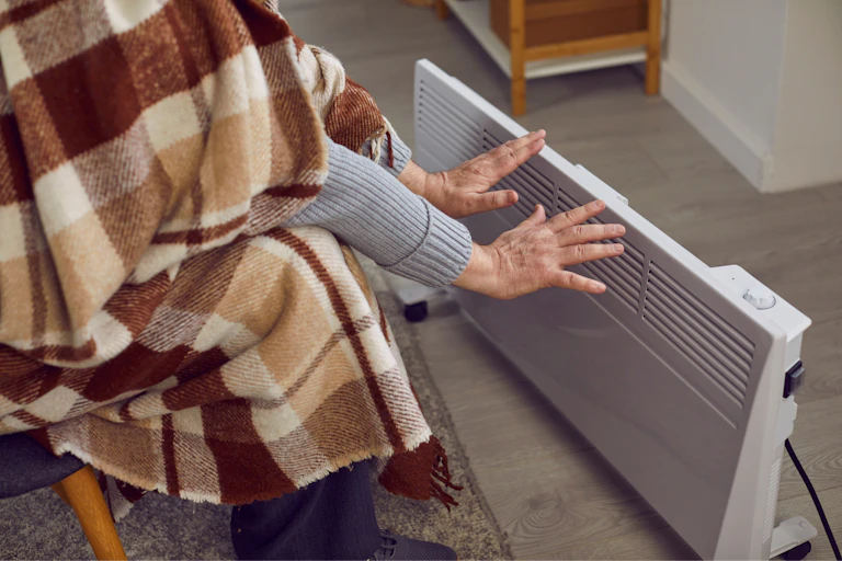 Une personne tâche de se réchauffer devant un petit radiateur d'appoint, avec une couverture sur le dos.
