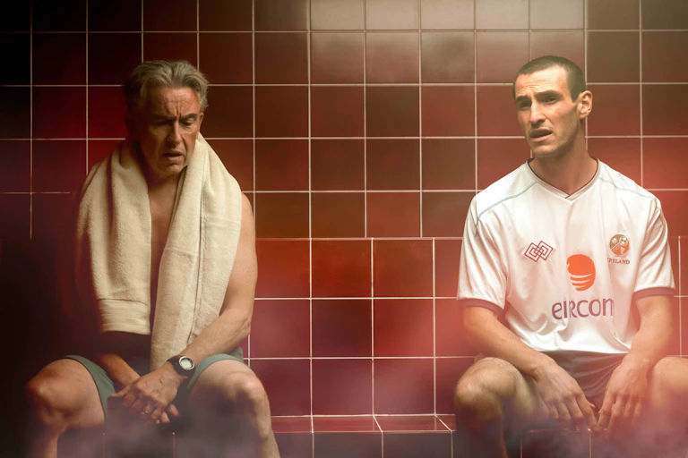 Two men, one older one in his early thirties, sitting in a tiled wet room talking,