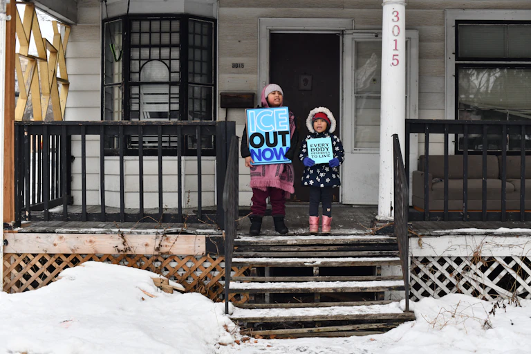 Two young children wear winter outerwear and hold signs that say 'ICE Out Now' and 'Everybody deserves to live.' They stand on the stoop of a house with a porch.