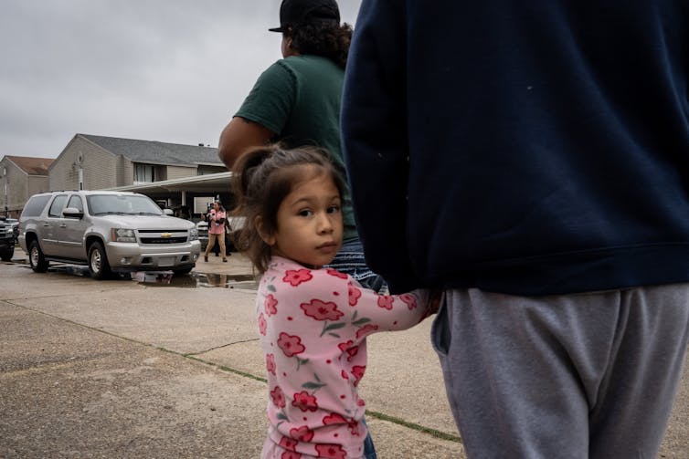 A young girl wearing a pink shirt holds an adult's hand and looks directly at the camera. She stands on a street near a parked gray SUV.