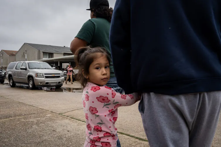 A young girl wearing a pink shirt holds an adult's hand and looks directly at the camera. She stands on a street near a parked gray SUV.