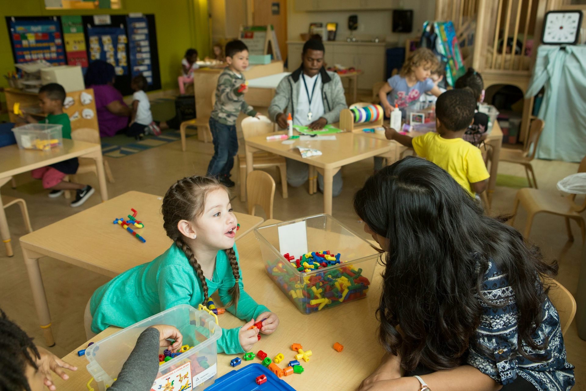 Young children with educators seen in a classroom at tables doing various activities with building blocks and other materials.