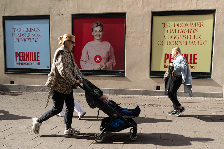 Uma mulher empurra um carrinho com um bebê passando por um cartaz eleitoral, enquanto outra mulher cruza seu caminho carregando objetos de forma desajeitada.