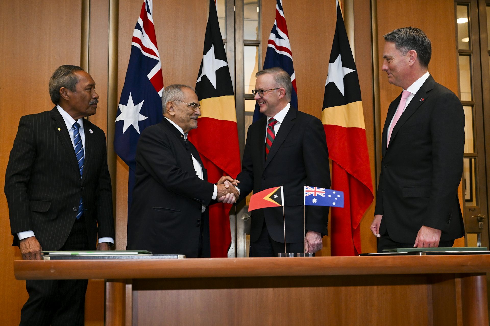 Four middle-aged men in suits stand in an office front of a set of country flags.