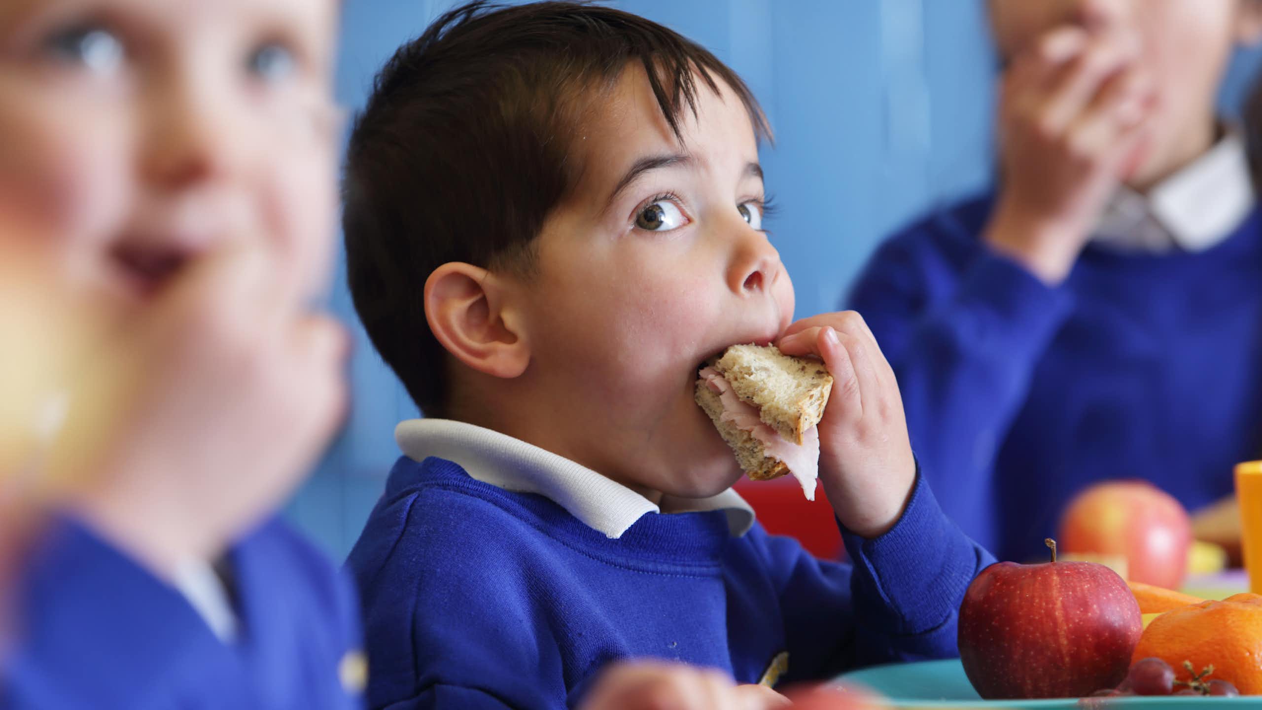 A young boy in school uniform puts a sandwich in his mouth.