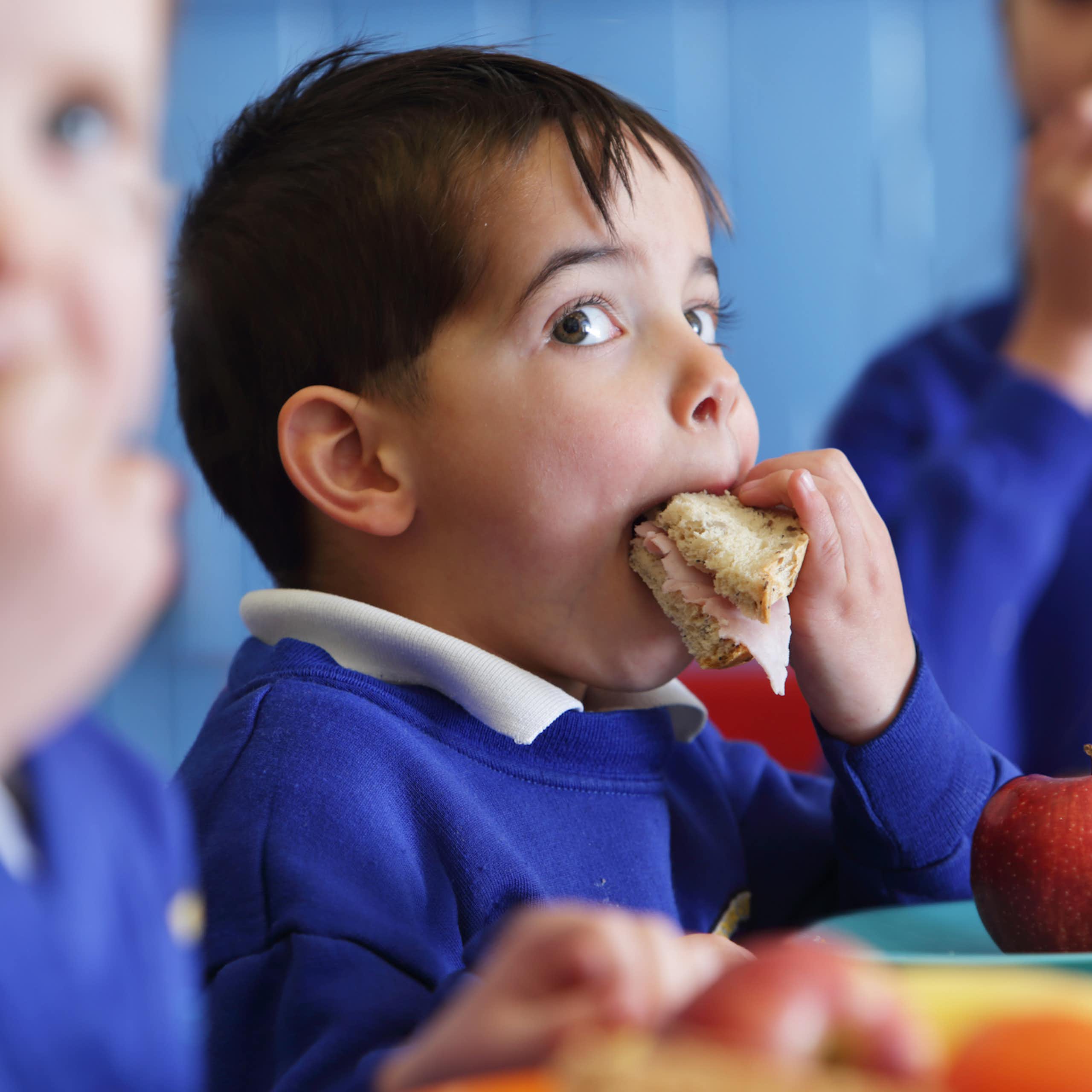 A young boy in school uniform puts a sandwich in his mouth.