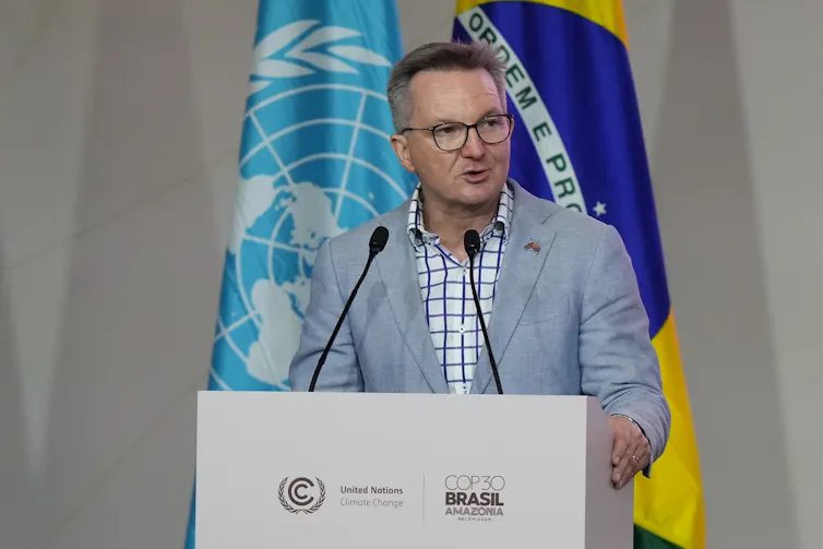 A middle-aged man in a grey suit delivers a speech while standing in front of the two flags.