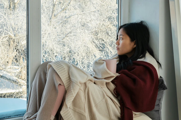 A woman wrapped in blankets and holding a mug sits on a windowsill