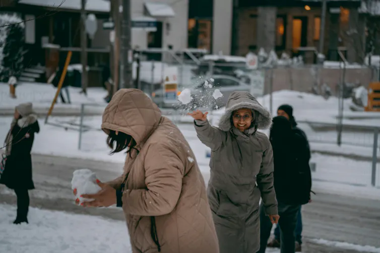 Two women in hooded winter coats tossing snow at each other on a city sidewalk