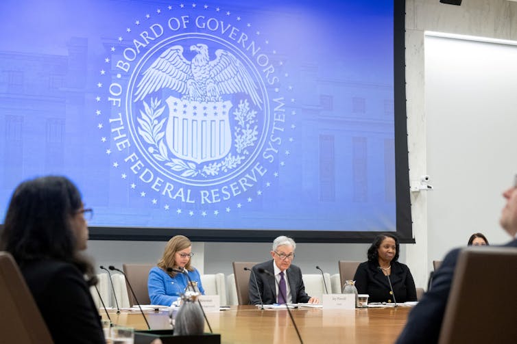 A group of people meet at a conference table while the Federal Reserve insignia is projected onto a screen above their heads.