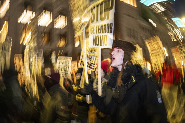A group of people hold signs that say 'Stop ICE's Terror' in a blurred photo with nighttime lights.