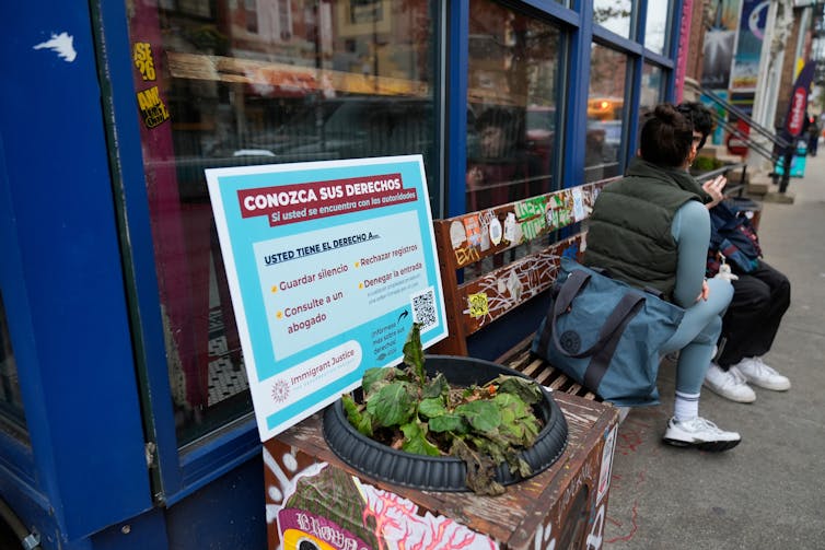 People sit on a bench near a sign in Spanish that says 'Conozca sus derechos'