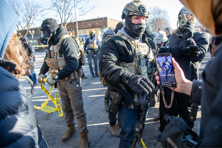 several men in tactical gear stand near several people holding up smartphones