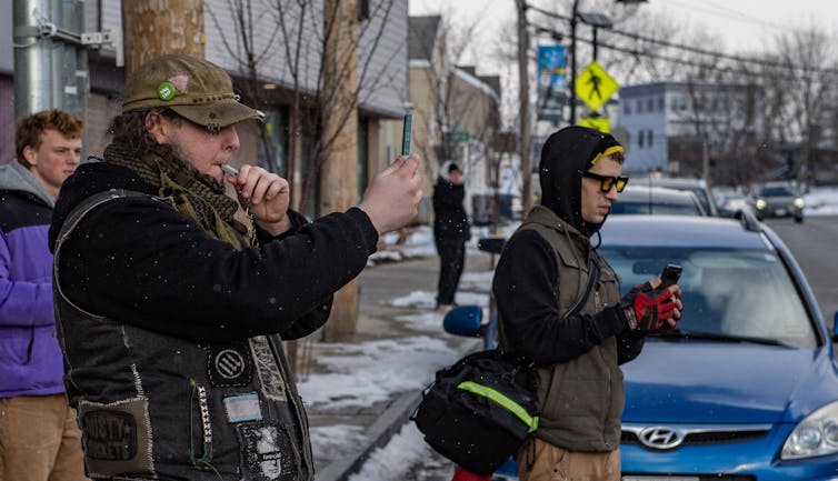 Two people on a sidewalk, one blowing a whistle and the other filming with a camera at something on the road.
