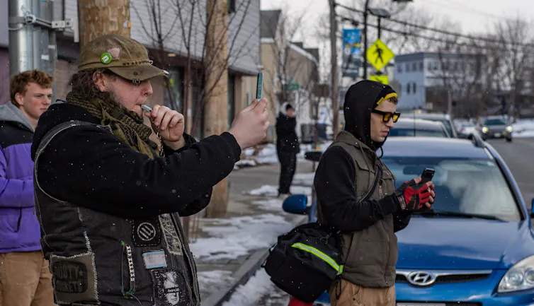Two people on a sidewalk, one blowing a whistle and the other filming with a camera at something on the road.