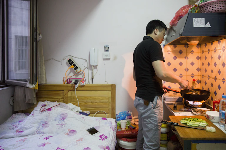 Man cooking at a stove in a small room next to a bed