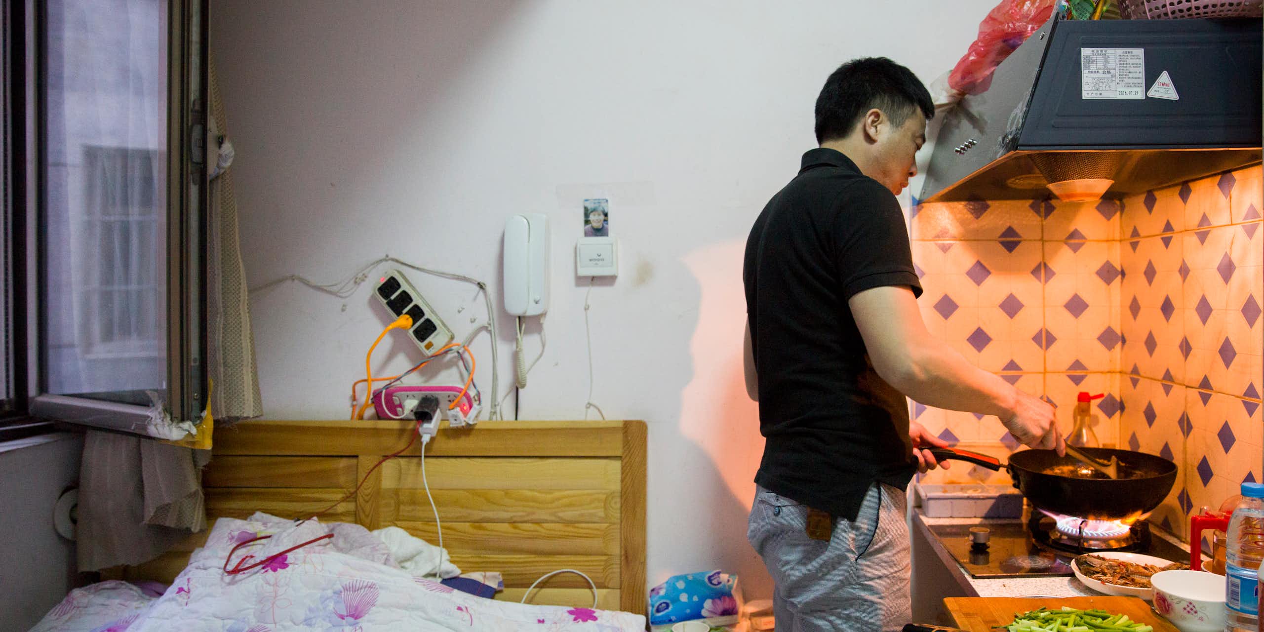 Man cooking at a stove in a small room next to a bed