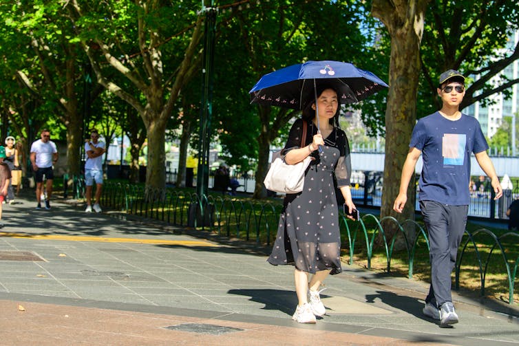 A woman, holding an umbrella, and a man walk along a city footpath in the sun.