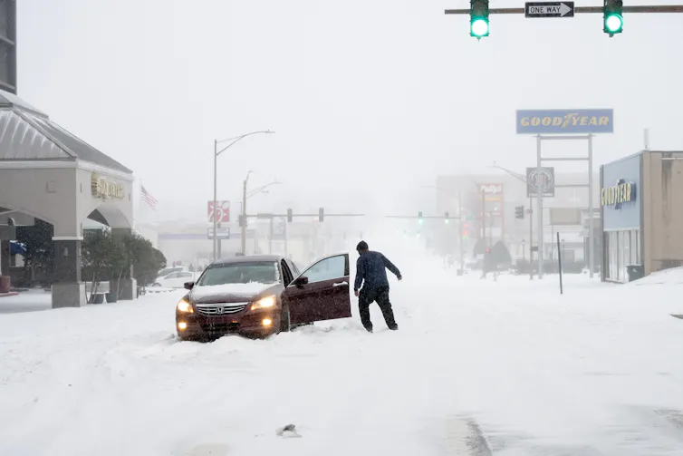 A man stands by the open door of a car stuck on a road with deep snow.