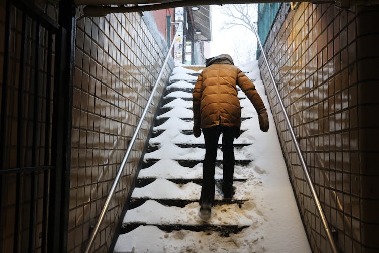 A woman in a puffy jacket, hat and scarf walks up snow-covered subway stairs.