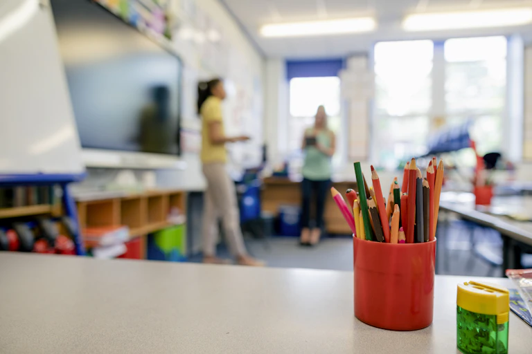 An empty school classroom, with a cup of coloured pencils. Two adults stand in the background.
