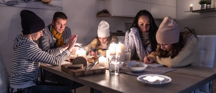 Five people sit around a table, each wrapped up in warm clothes and hats. Two children are studying.