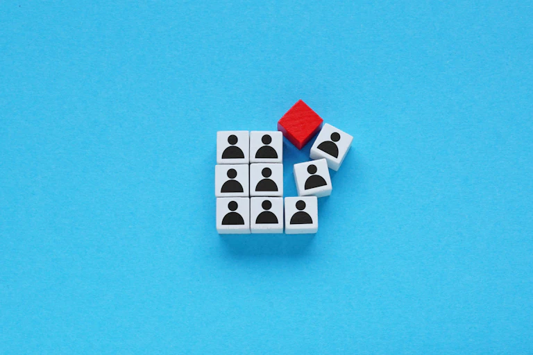 A neat square of dice like objects with human silhouettes on them broken by one random red cube smashing into them.