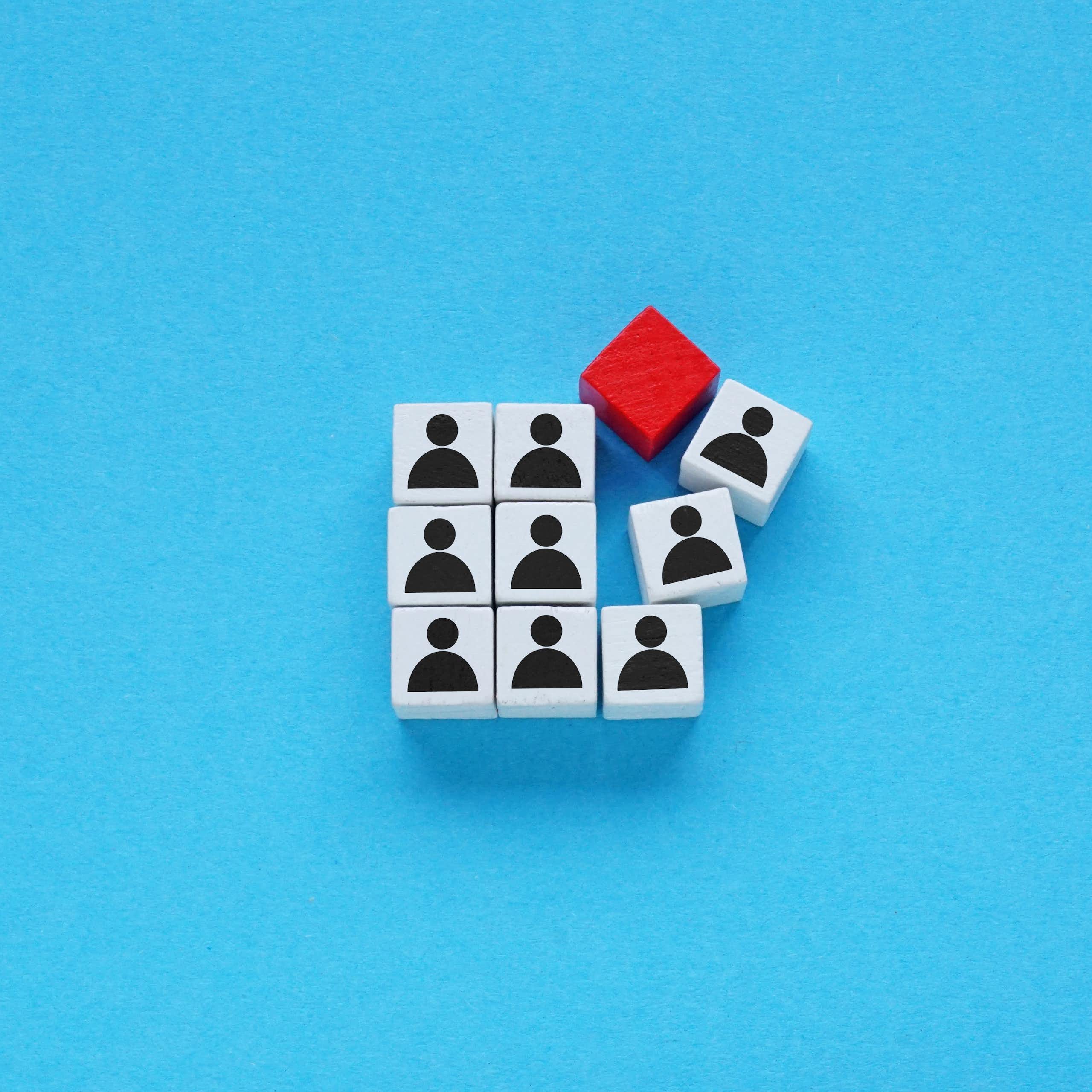A neat square of dice like objects with human silhouettes on them broken by one random red cube smashing into them.
