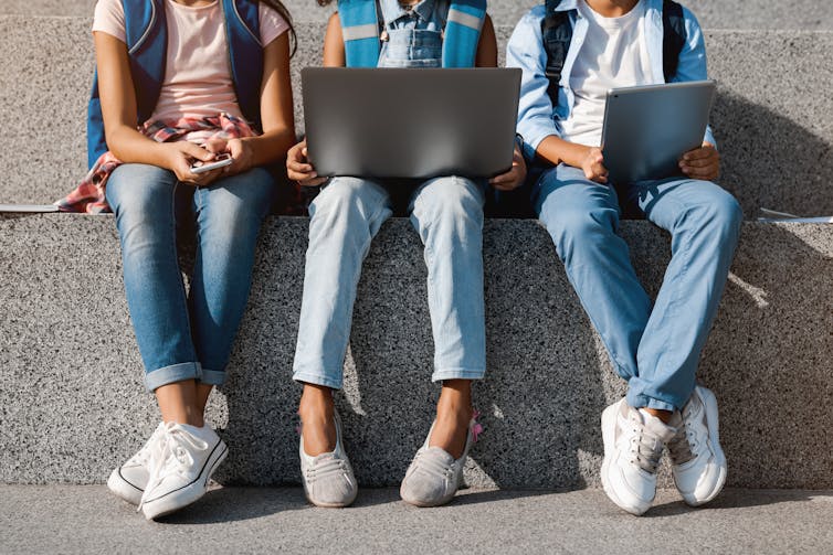 cropped photo of three young people from the neck down, sitting on a concrete step and looking at computers and tablets