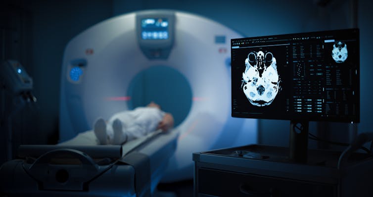 A person lays inside a PET scan machine. A screen to the right shows the ongoing scan of their brain.