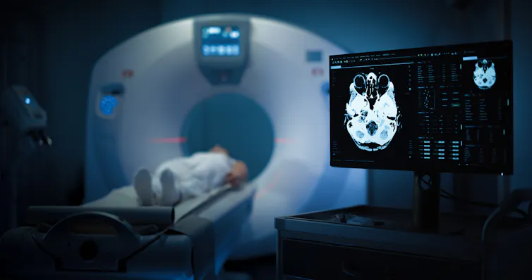 A person lays inside a PET scan machine. A screen to the right shows the ongoing scan of their brain.
