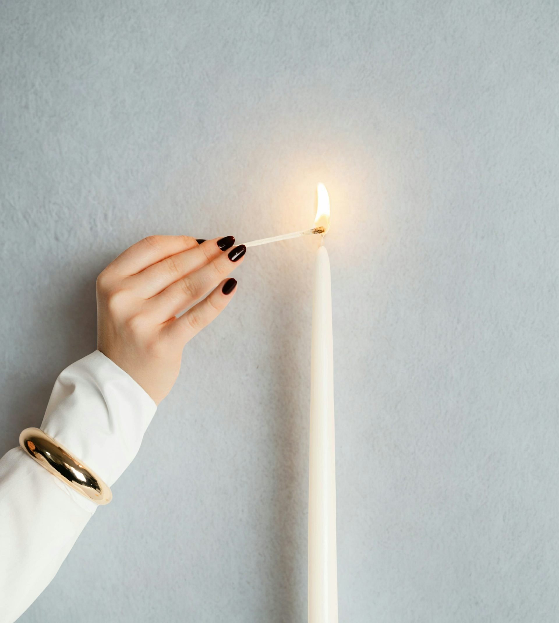Woman wearing white jumper with black painted nails lights a white candle with a match.