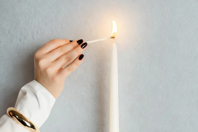 Woman wearing white jumper with black painted nails lights a white candle with a match.