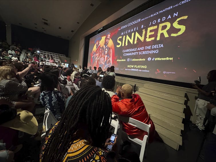 Una audiencia mayoritariamente negra se sienta y escucha a un panel de oradores negros en discusión bajo una pantalla grande que muestra un cartel de película que dice