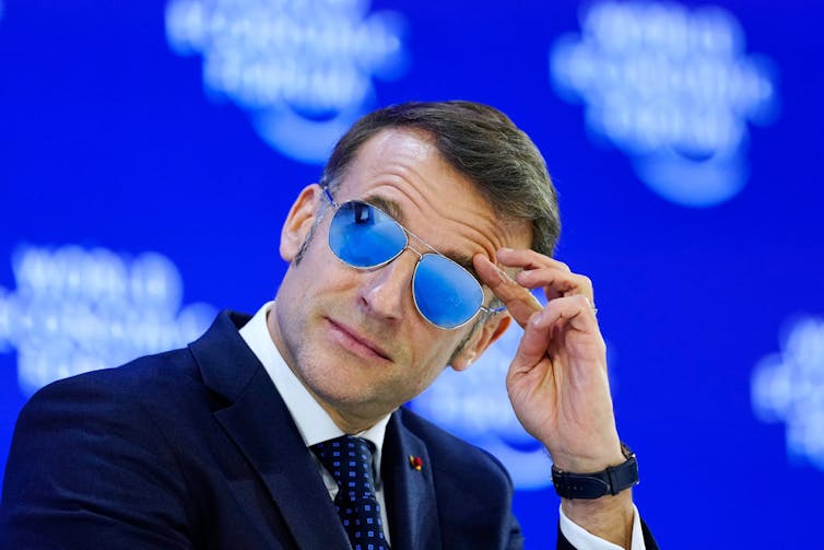 A man in sunglasses and a suit touches his head.