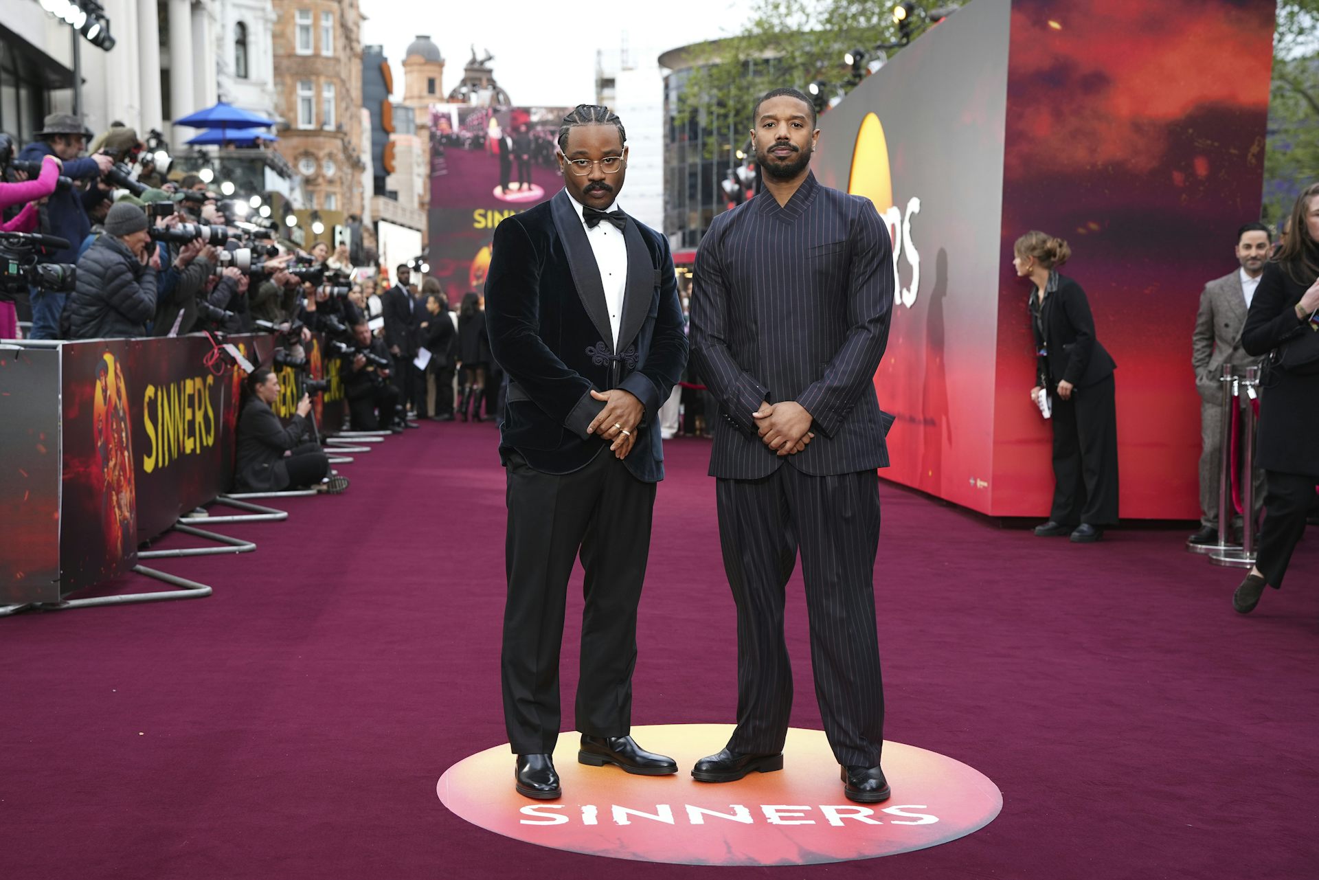 Dos hombres negros vestidos de forma formal se encuentran en la alfombra roja junto a fotógrafos de prensa.