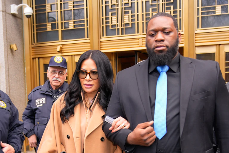 A woman with long dark hair and a beige coat holds the arm of a tall man wearing a black suit and blue tie and walks away from a building, with police nearby.