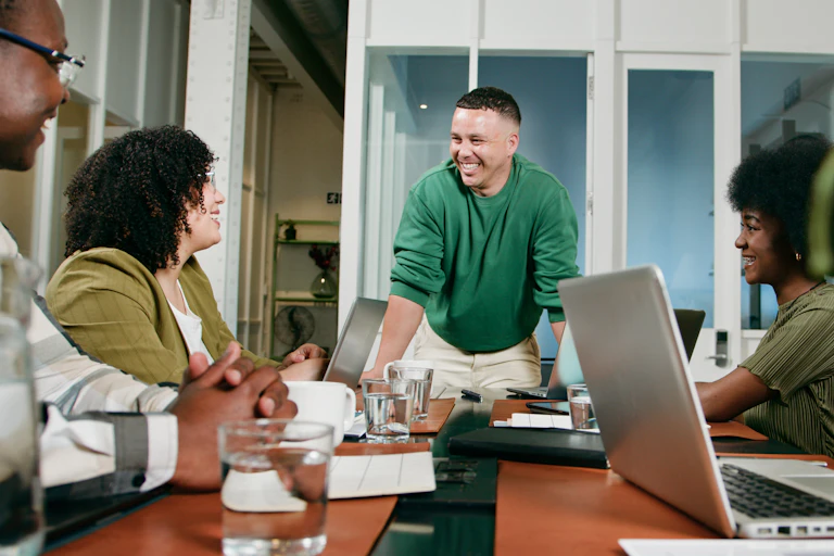 man stands at head of a work table with seated people looking at him, all smiling