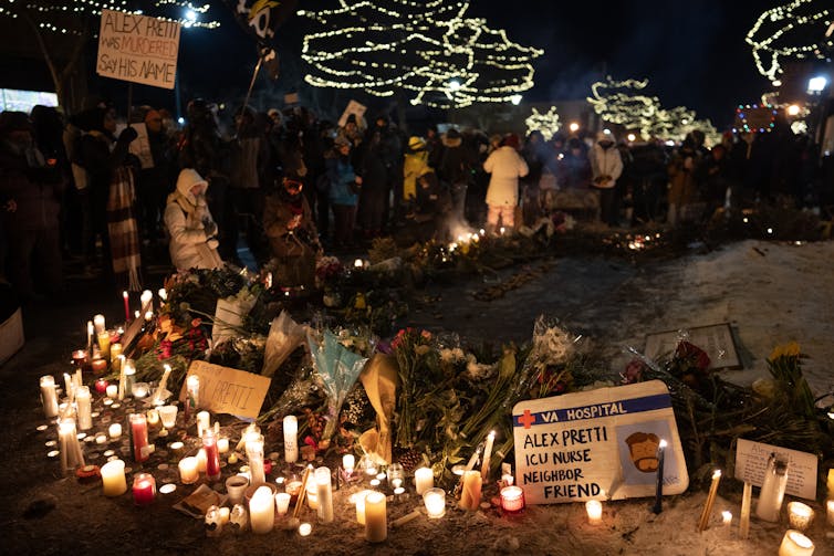 Many people in the nighttime standing next to a memorial of candles and signs about the killing of Alex Pretti.