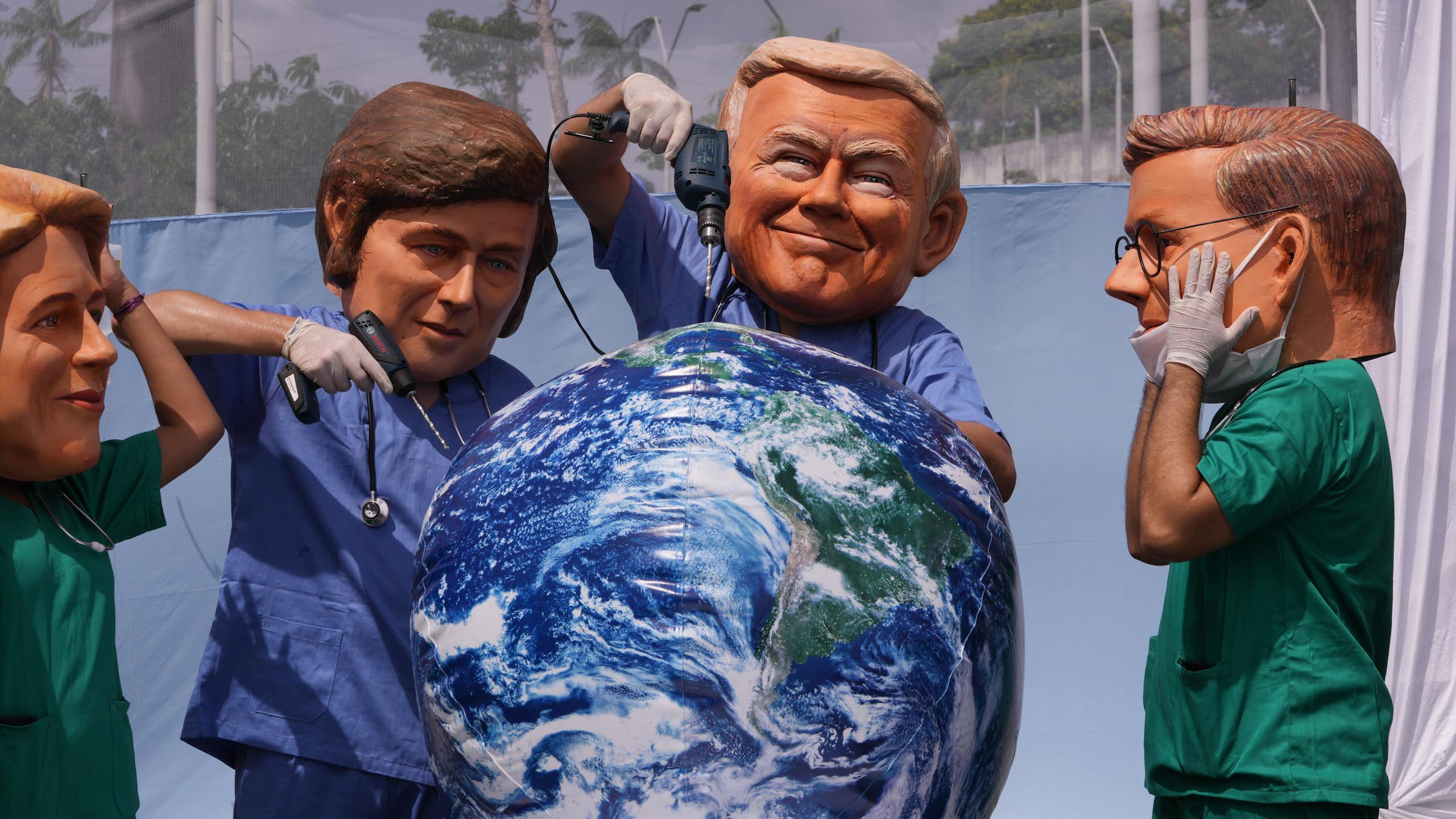 People wearing large masks bearing the likeness of donald trump and javier milei drill into a display of the planet with power drills