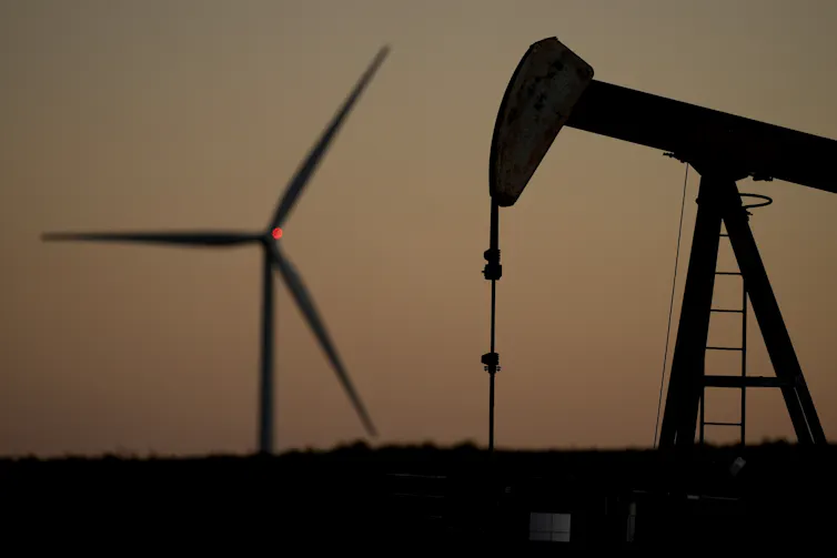 A pumpjack operates in the foreground while a wind turbine is seen in the background at sunset